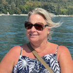 Woman in sunglasses smiling on a boat with water and greenery behind her.